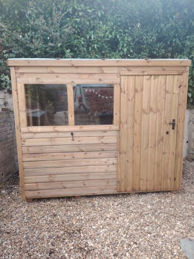 Wooden shed with a window, located in a gravel area next to greenery.