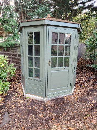 A small green garden shed with multiple windows and a door, situated among greenery.